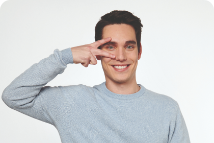hombre joven con jersey gris haciendo la señal de la victoria sobre un ojo